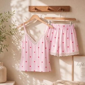 May include: Pink pajama set with red heart pattern. The set includes a camisole top with lace trim and matching shorts. The camisole has thin straps and a V-neck. The shorts have a drawstring waist and lace trim at the hem.