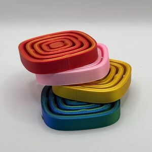 May include: A stack of four square-shaped coasters in red, pink, yellow, and blue. The coasters are made of plastic and have a concentric circle design.