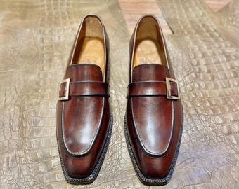 Pode incluir: Um par de mocassins de couro castanho com detalhe de fivela. Os sapatos têm um acabamento polido e uma biqueira quadrada. Os mocassins são exibidos em uma superfície texturizada marrom claro.