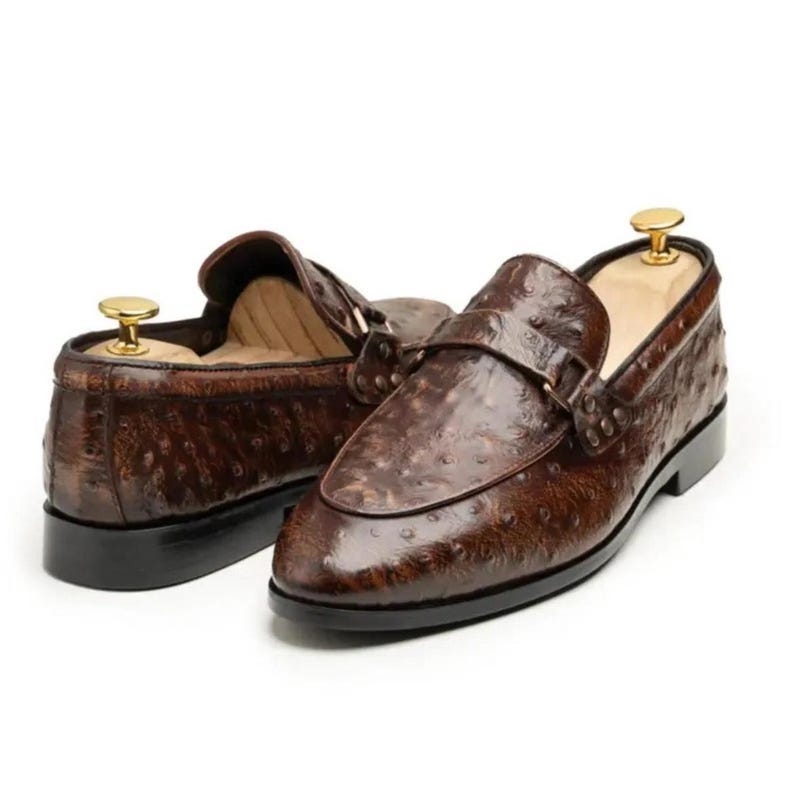Pu&ograve; includere: Un paio di mocassini in pelle marrone con una superficie strutturata e fantasia. Le scarpe hanno un design classico con un cinturino sulla parte superiore e una suola nera. Le forme sono inserite all'interno.