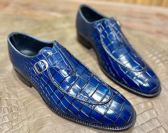 May include: A pair of blue leather dress shoes with a crocodile skin pattern. The shoes feature a buckle closure and a glossy finish. The shoes are displayed on a textured surface.