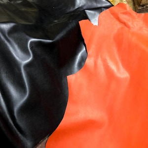May include: Two pieces of leather are shown, one black and one bright orange. The black leather has a textured surface with subtle vertical lines. The orange leather is smooth and vibrant. The leather pieces are irregularly shaped, suggesting they are cut from larger hides.