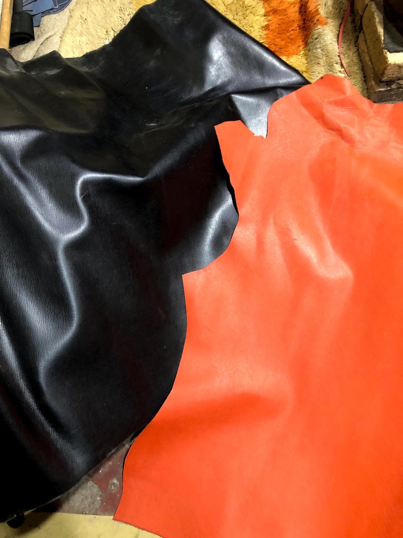 May include: Two pieces of leather are shown, one black and one bright orange. The black leather has a textured surface with subtle vertical lines. The orange leather is smooth and vibrant. The leather pieces are irregularly shaped, suggesting they are cut from larger hides.