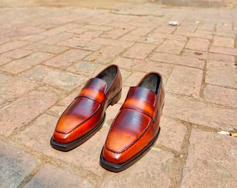 Pode incluir: Um par de mocassins de couro castanho com um efeito gradiente, passando de um castanho escuro para um tom mais claro. Os sapatos têm um design clássico slip-on com bico quadrado e sola preta. O couro parece polido.