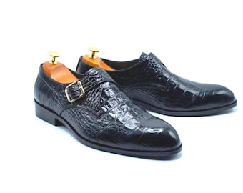 May include: A pair of black leather dress shoes with a crocodile skin texture. The shoes feature a single buckle closure with a gold-colored metal buckle. Wooden shoe trees are inserted inside the shoes.