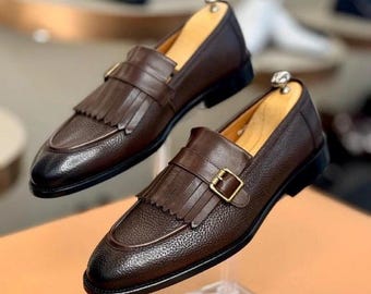Pode incluir: Um par de mocassins de couro castanho com acabamento texturizado. Cada sapato apresenta uma tira com fivela dourada e franjas decorativas. Os sapatos são exibidos em suportes transparentes.