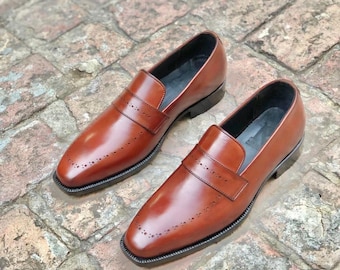 Pode incluir: Um par de mocassins de couro castanho. Os sapatos têm um acabamento polido, perfurações decorativas e sola preta. Os mocassins são exibidos numa superfície de tijolo.