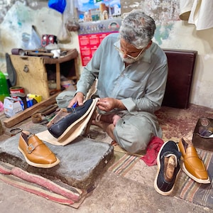 May include: A shoemaker at work, seated on the floor, crafting leather shoes. The image shows various stages of shoemaking, with finished and unfinished shoes in tan and black leather. Tools and materials are scattered around the workspace.