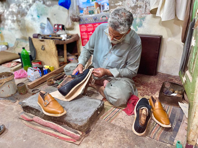May include: A shoemaker at work, seated on the floor, crafting leather shoes. The image shows various stages of shoemaking, with finished and unfinished shoes in tan and black leather. Tools and materials are scattered around the workspace.