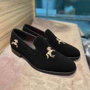 May include: Black slip-on shoes with gold horse embroidery. The shoes have a rounded toe and a low heel. The horse design is on the vamp and outer side of each shoe. The shoes are displayed on a neutral-toned surface.