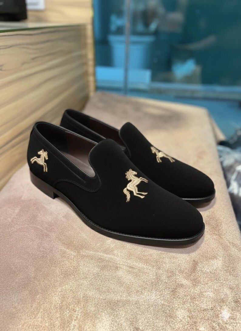 May include: Black slip-on shoes with gold horse embroidery. The shoes have a rounded toe and a low heel. The horse design is on the vamp and outer side of each shoe. The shoes are displayed on a neutral-toned surface.