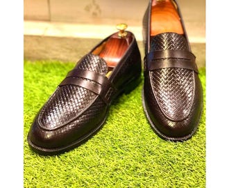Pode incluir: Um par de mocassins de couro castanho escuro com um padrão tecido na gáspea e uma tira na parte superior. Os sapatos são exibidos em uma superfície verde, mostrando seu design clássico e artesanato.