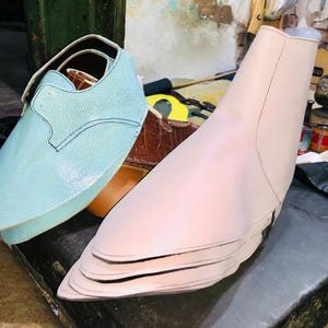 Pu&ograve; includere: Una scarpa in pelle azzurra con cinturino e dettagli di cucitura &egrave; accanto a una forma per scarpe rosa chiaro. Una scarpa in pelle marrone &egrave; parzialmente visibile sullo sfondo. Gli oggetti sono su una superficie scura.