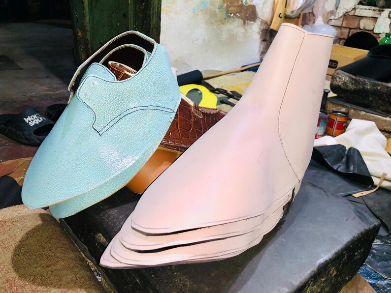 Pu&ograve; includere: Una scarpa in pelle azzurra con cinturino e dettagli di cucitura &egrave; accanto a una forma per scarpe rosa chiaro. Una scarpa in pelle marrone &egrave; parzialmente visibile sullo sfondo. Gli oggetti sono su una superficie scura.