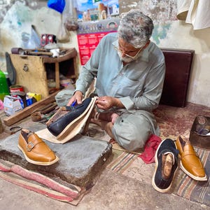 May include: A craftsman seated on the floor, making leather shoes. The image shows the shoemaker working on a black leather shoe, with other finished and unfinished shoes in tan and black. Tools and materials are visible in the workshop.