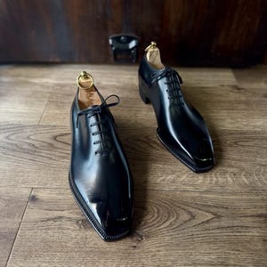 以下が含まれることがあります： 黒革のレースアップシューズのペア。つま先が尖っていて、かかとが四角い。靴は木製の表面の上に置かれている。