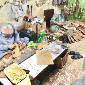 Pu&ograve; includere: Due uomini sono mostrati mentre lavorano su calzature in pelle in un laboratorio. Sono seduti sul pavimento, circondati da strumenti, materiali e scarpe parzialmente completate. L'ambiente sembra essere un ambiente tradizionale di calzoleria.