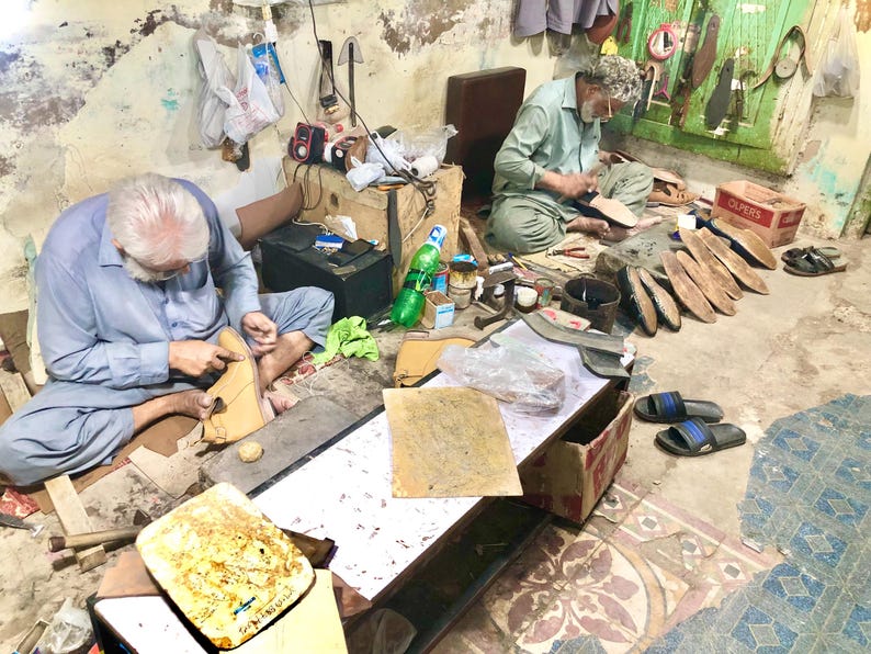 Pu&ograve; includere: Due uomini sono mostrati mentre lavorano su calzature in pelle in un laboratorio. Sono seduti sul pavimento, circondati da strumenti, materiali e scarpe parzialmente completate. L'ambiente sembra essere un ambiente tradizionale di calzoleria.