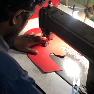 May include: A person using a vintage sewing machine to stitch red material. The machine is black and branded "SEASTAR". Blue scissors lie on the wooden work surface. The scene suggests a workshop or manufacturing setting.