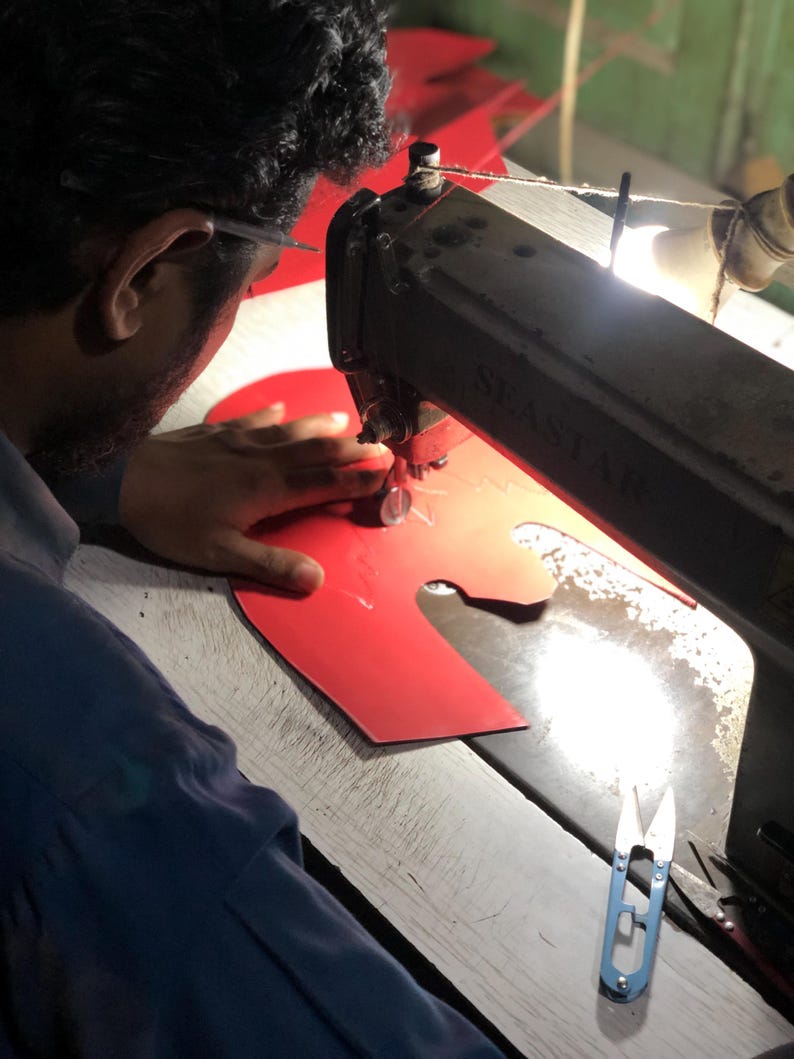 May include: A person using a vintage sewing machine to stitch red material. The machine is black and branded "SEASTAR". Blue scissors lie on the wooden work surface. The scene suggests a workshop or manufacturing setting.
