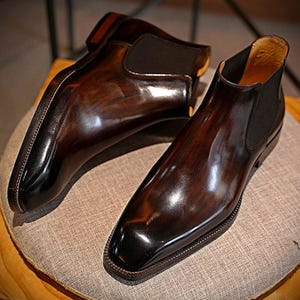 May include: A pair of polished, brown leather Chelsea boots. The boots have a glossy finish with a dark brown hue and a square toe. The elastic side panels are black. The boots are placed on a neutral-colored, textured surface.