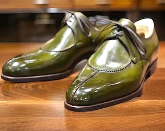 May include: A pair of green leather dress shoes with a glossy finish. The shoes have a classic design with laces and a stitched seam down the center of the vamp. The soles are dark brown, and the shoes are set on a wooden surface.