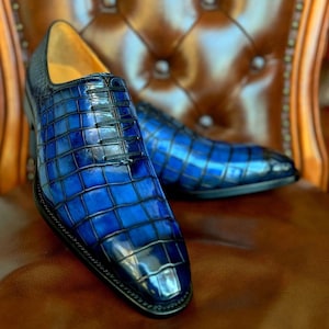 May include: A pair of blue leather dress shoes with a crocodile skin pattern. The shoes have black laces and a black sole. The shoes are on a brown leather chair.