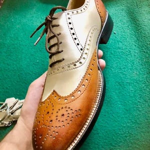 May include: A two-tone leather oxford shoe with brown laces. The shoe features a cream-coloured vamp and quarter, with brown detailing on the toe cap and heel. The shoe has decorative perforations and a black sole.