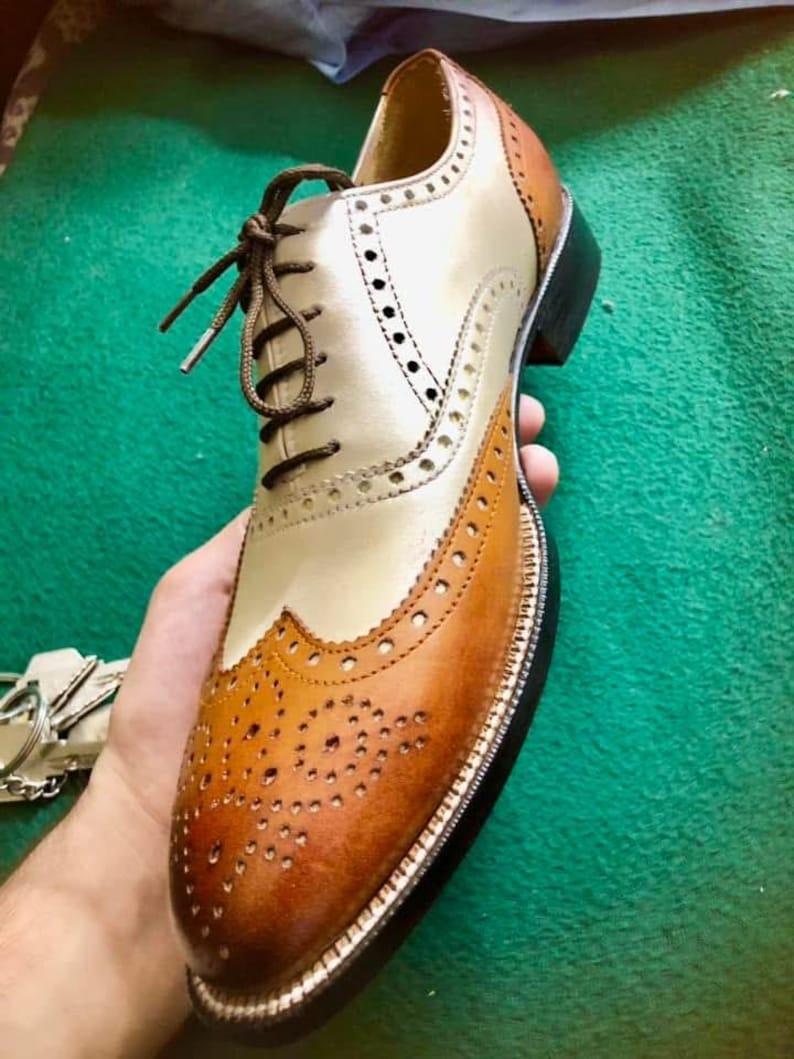 May include: A two-tone leather oxford shoe with brown laces. The shoe features a cream-coloured vamp and quarter, with brown detailing on the toe cap and heel. The shoe has decorative perforations and a black sole.