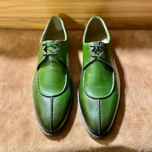 Puede incluir: Un par de zapatos de cuero verde oscuro con un diseño de punta dividida y una costura vertical prominente que recorre el empeine. Los zapatos cuentan con un cierre de cordones finos negros y un acabado de cuero liso y pulido.