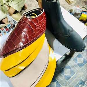 May include: A partially constructed shoe with a burgundy crocodile-textured leather upper and a stacked sole with yellow and beige layers. A black leather boot with decorative perforations is also present.