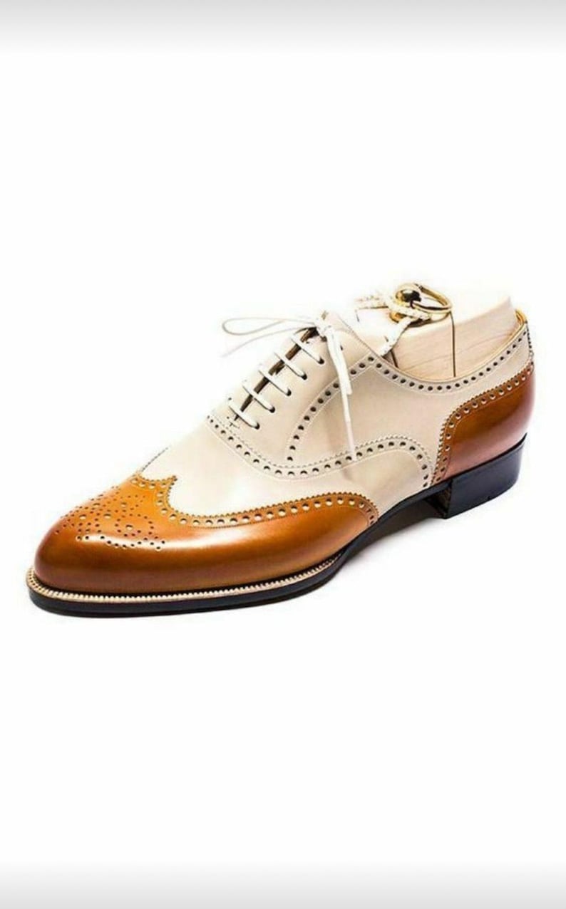 May include: Two-tone leather oxford shoe in cream and brown. The shoe features a wingtip design with decorative perforations and a classic lace-up closure. The sole is black with a thin white trim.