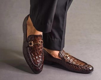 May include: Brown leather loafers with a crocodile texture and gold-colored buckle detail. The shoes are a dark brown color and appear to be new. The loafers are a classic style of footwear.