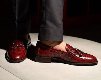 Pode incluir: Mocassins de couro vermelho com padrão de crocodilo. Os sapatos têm borlas e um salto baixo. A pessoa está usando calças cinza. Os sapatos têm cerca de 25 cm de comprimento.