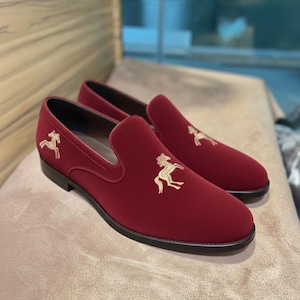 May include: A pair of burgundy slip-on loafers with a gold embroidered horse emblem on the vamp. The shoes have a rounded toe and a black sole. The loafers are made of a soft, velvety material.