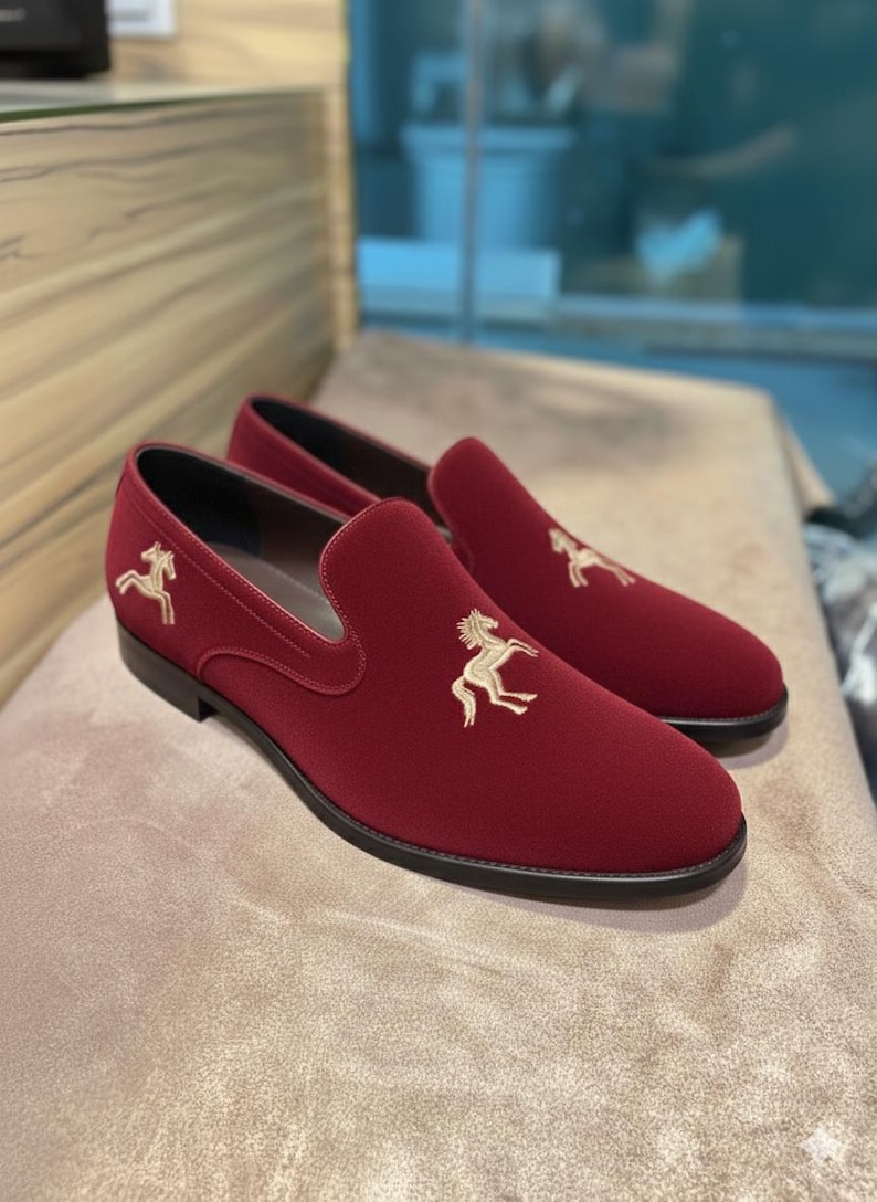 May include: A pair of burgundy slip-on loafers with a gold embroidered horse emblem on the vamp. The shoes have a rounded toe and a black sole. The loafers are made of a soft, velvety material.