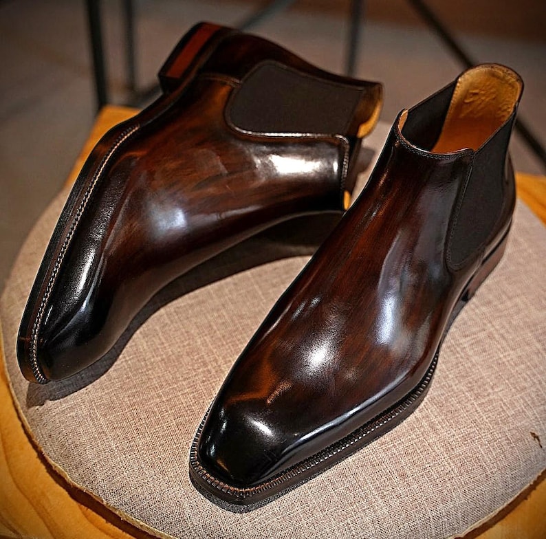 May include: A pair of polished, brown leather Chelsea boots with a glossy finish. The boots have a classic design with elastic side panels and a square toe. The leather has a rich, multi-tonal appearance, with darker shades at the toe and heel.