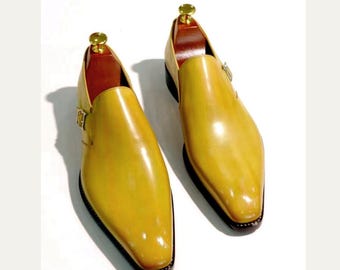 May include: A pair of yellow leather dress shoes with a glossy finish. The shoes have a pointed toe, a buckle closure, and a dark sole. Shoe trees are inserted inside the shoes. The shoes are displayed on a white background.