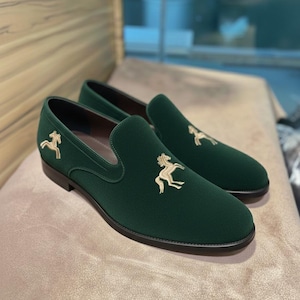 May include: A pair of dark green velvet loafers with a gold embroidered horse emblem on the vamp. The slip-on shoes have a rounded toe and a black sole. The shoes are displayed on a neutral-toned surface.
