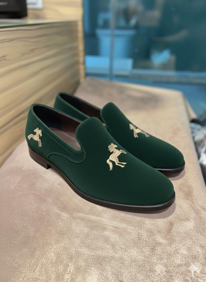May include: A pair of dark green velvet loafers with a gold embroidered horse emblem on the vamp. The slip-on shoes have a rounded toe and a black sole. The shoes are displayed on a neutral-toned surface.