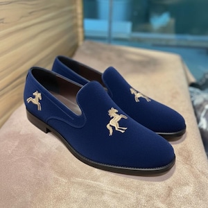 May include: A pair of navy blue slip-on loafers with gold horse embroidery. The shoes have a rounded toe and a low heel. The loafers are made of a soft, velvety material. The shoes are displayed on a neutral-colored surface.