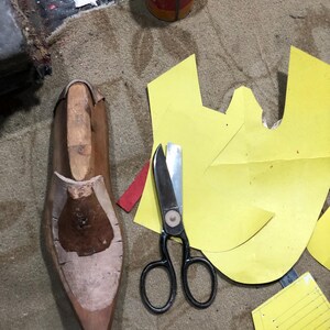 May include: A shoemaking workshop scene with a wooden shoe last, black scissors, and yellow paper patterns. The tools are arranged on a textured, brown surface, suggesting a craft or artisan setting.