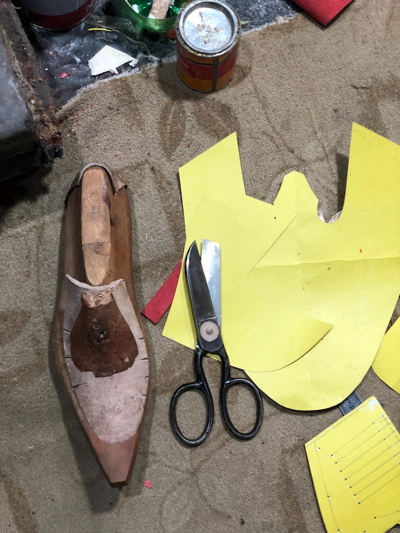 May include: A shoemaking workshop scene with a wooden shoe last, black scissors, and yellow paper patterns. The tools are arranged on a textured, brown surface, suggesting a craft or artisan setting.