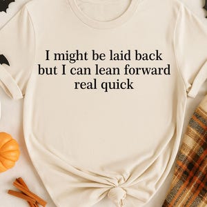 May include: Cream-colored t-shirt with the text "I might be laid back but I can lean forward real quick" in black. The shirt is styled with Halloween decorations, including bats, a ghost, a pumpkin, and cinnamon sticks.