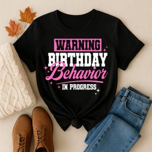 May include: A black short-sleeved t-shirt featuring the white and pink text WARNING BIRTHDAY BEHAVIOR IN PROGRESS. The design includes small star accents and is centered on the chest of the garment, which has a crew neckline and a knotted hem.