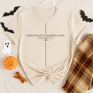 May include: A cream-colored short-sleeve t-shirt featuring a black vertical line forming a cross shape. The text reads "fatherhood is kingdom work" followed by "PROVERBS 22:6" and a small crown icon centered on the front of the garment.