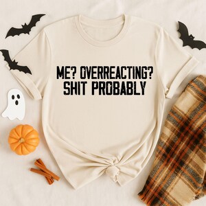 May include: A cream-colored short-sleeve t-shirt featuring the black text ME? OVERREACTING? SHIT PROBABLY printed in a distressed font across the chest. The shirt has a crew neckline and is made of a solid, light-colored fabric with a simple design.