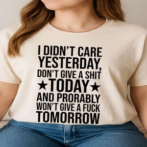 Könnte beinhalten: Cremefarbenes T-Shirt mit schwarzem Text: "I DIDN'T CARE YESTERDAY, DON'T GIVE A SHIT TODAY AND PROBABLY WON'T GIVE A FUCK TOMORROW", mit Sternsymbolen. Das Shirt wird mit einer blauen Jeans getragen.