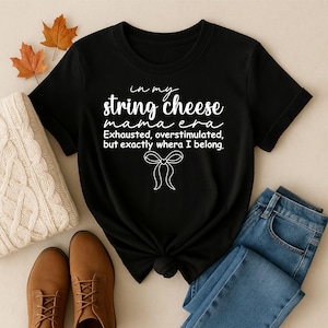 Könnte beinhalten: Schwarzes T-Shirt mit dem Text "in my string cheese mama era Exhausted, overstimulated, but exactly where I belong." Das Shirt ist mit blauen Jeans, braunen Stiefeln und einem cremefarbenen Pullover gestylt.