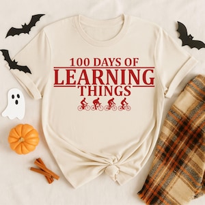 May include: A cream-coloured t-shirt featuring the text "100 Days of Learning Things" in red, and four cyclists. The image includes Halloween decorations such as bats, a ghost, a pumpkin, and cinnamon sticks.